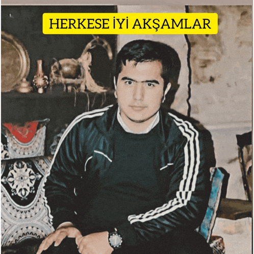 a man in a black jacket sits in a chair under a yellow sign that says herkese iyi aksamlar