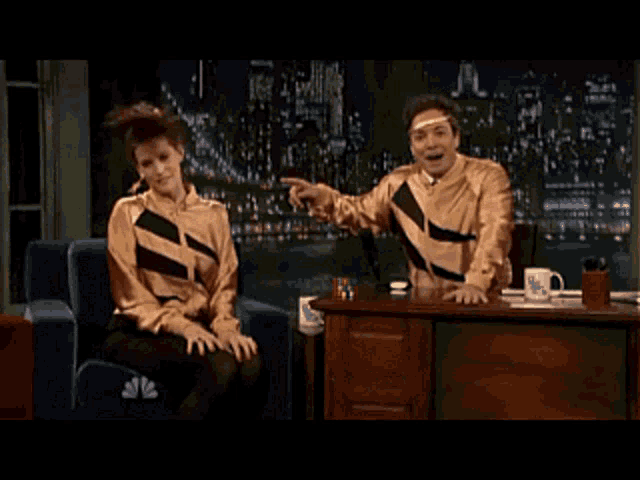 a man and a woman are sitting at a desk in front of a nbc mug