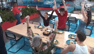 a group of people are sitting at a table with plates of food and raising their arms in the air .