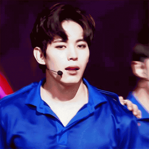 a young man wearing a blue shirt and a microphone looks at the camera