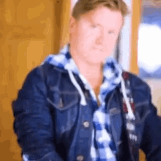 a man wearing a denim jacket and a plaid shirt is sitting in front of a window .