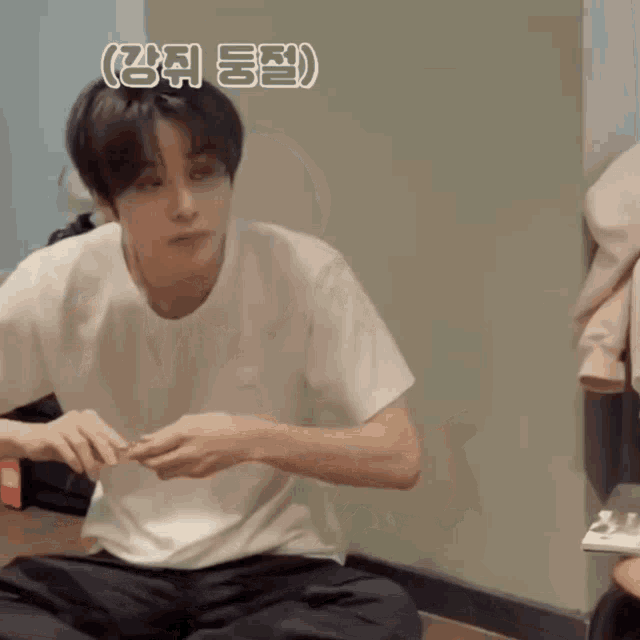a young man in a white t-shirt is sitting on the floor with his hands folded