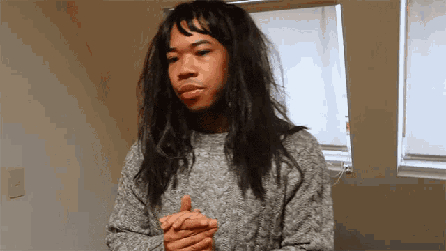 a man with long hair wearing a grey sweater stands in front of a window