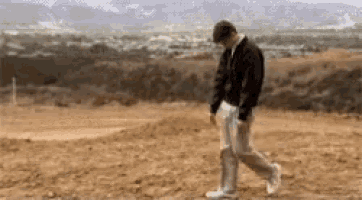 a man in a black jacket and white pants is walking through a dirt field .