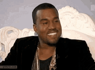 a man in a black jacket and gold chains is smiling while sitting on a white couch .