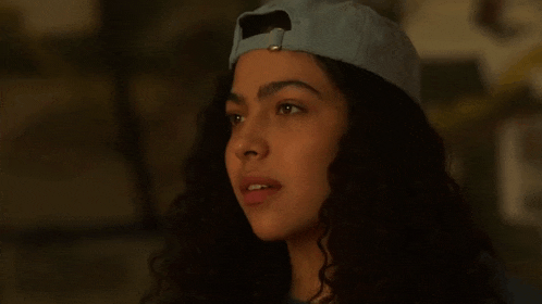 a young woman with curly hair is wearing a baseball cap and looking at the camera .