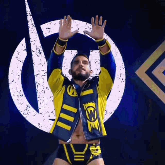 a wrestler in a yellow and blue jacket is standing in front of a logo with his hands in the air .