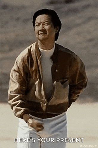 a man in a brown jacket and white shorts is standing in a field and says here 's your present .
