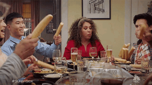 a group of people are sitting around a table eating food and drinking wine while holding bread sticks in the air ..