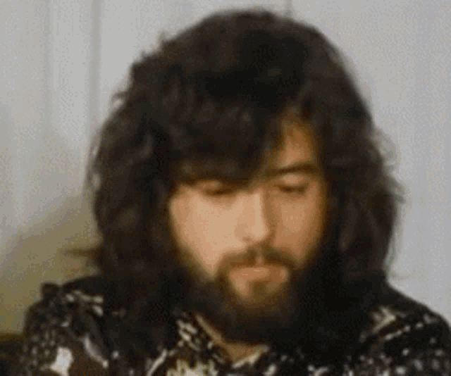 a man with long hair and a beard is sitting in front of a white wall .