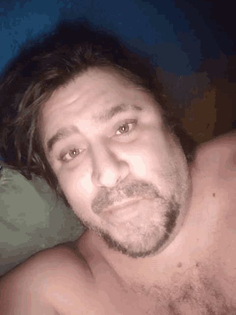 a shirtless man with a beard and long hair is laying in bed looking at the camera .