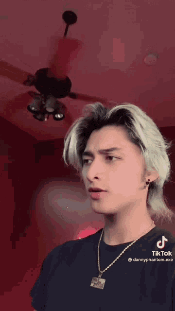a man wearing a black shirt and a gold necklace is standing in front of a ceiling fan in a room .