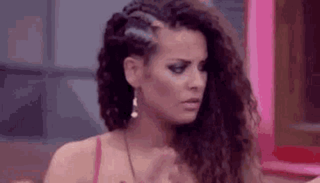 a woman with long curly hair is wearing a red top and earrings and making a funny face .