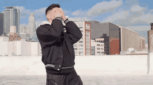 a man in a black jacket is covering his face with his hands in front of a city skyline