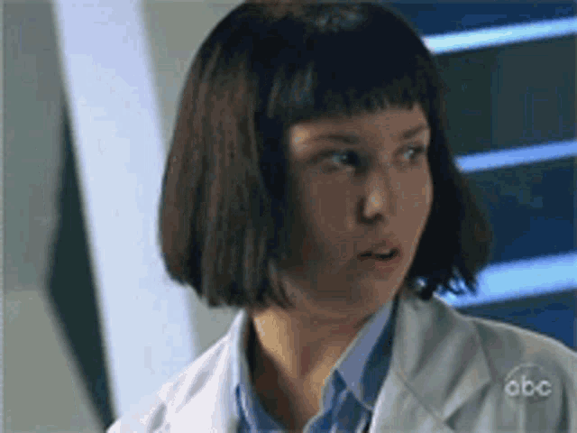 a woman wearing a white lab coat and a blue shirt is looking at the camera .