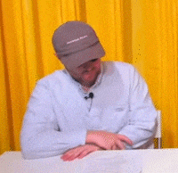 a man wearing a hat and sunglasses sits at a table in front of a yellow curtain