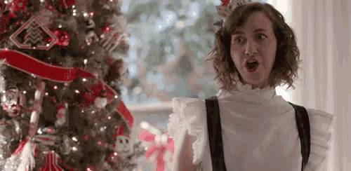 a woman in a maid costume is standing in front of a christmas tree and making a surprised face .