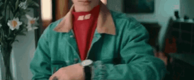 a man wearing a green jacket and a red turtleneck is sitting at a table looking at his watch .