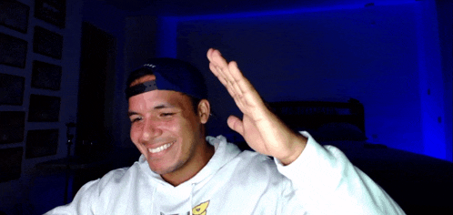 a man wearing a white hoodie and a blue hat is smiling and waving
