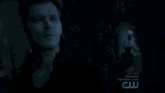 a man in a black shirt is standing in a dark room with a cw logo behind him .