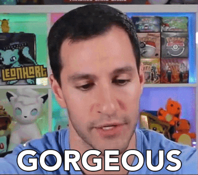 a man in a blue shirt is standing in front of a shelf with pokemon cards and toys and says gorgeous .