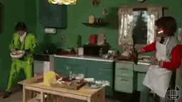a man in green pants is standing in a kitchen while a woman in an apron is cooking .
