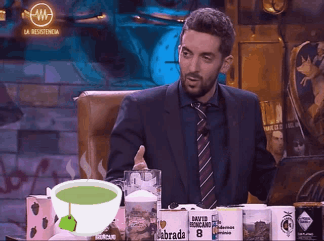 a man in a suit and tie sits in front of a table full of mugs one of which says " la resistencia "