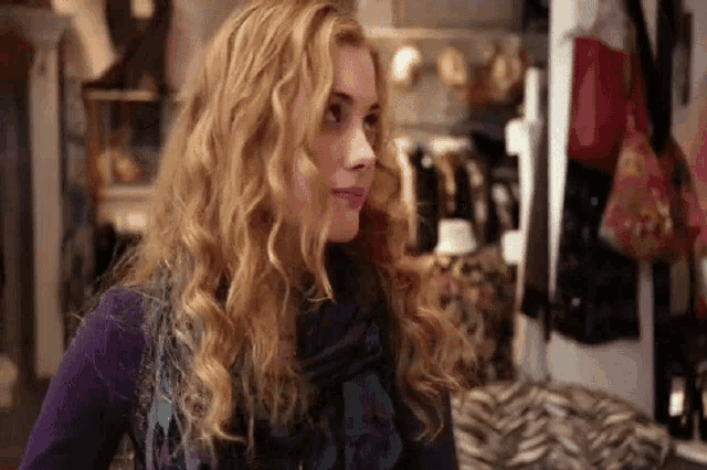 a woman with blonde curly hair is wearing a scarf and a purple sweater in a clothing store .