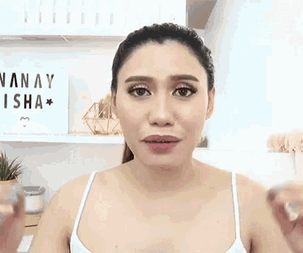 a woman in a white tank top is making a funny face in front of a sign that says vanay isha .