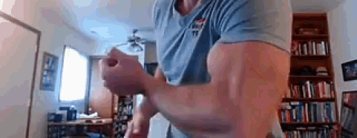 a man is flexing his muscles in front of a bookshelf in a living room .