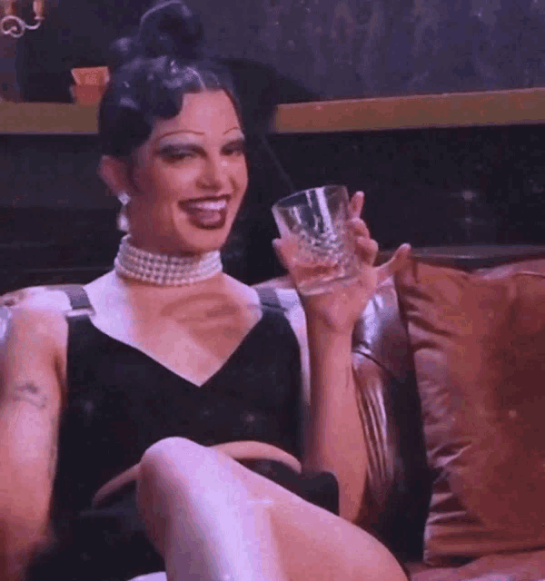 a drag queen is sitting on a couch holding a glass