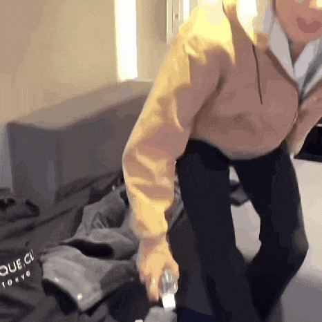 a man in a yellow jacket is standing on a bed holding a bottle .
