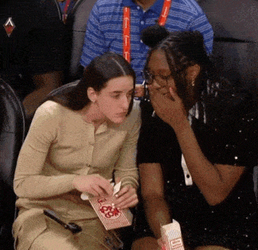 two women are sitting in a stadium eating popcorn and one is covering her mouth with her hands