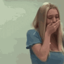 a blonde woman in a blue shirt is covering her mouth with her hand .
