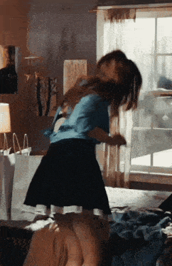 a woman in a blue shirt and black skirt is standing on a bed in front of a window