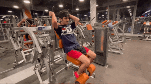 a man in a fila shirt sits on an orange machine in a gym