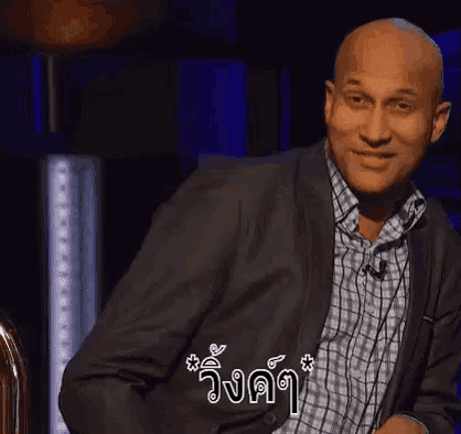 a bald man in a suit and plaid shirt is smiling while sitting in a chair .