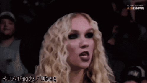 a blonde woman with curly hair is sitting in a crowd watching a wrestling match sponsored by jungle hooks / tumblr