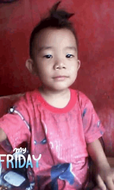 a young boy with a mohawk is wearing a red shirt and sitting in front of a red wall .