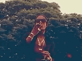 a man wearing sunglasses and a necklace is smoking a cigarette in front of a forest .