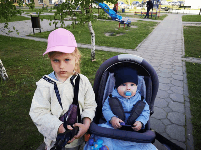 a little girl wearing a pink hat stands next to a baby