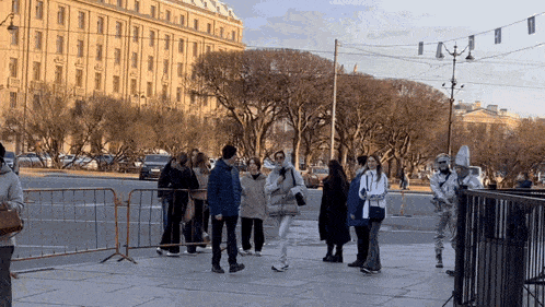 a group of people are standing on a sidewalk in front of a building with a fence