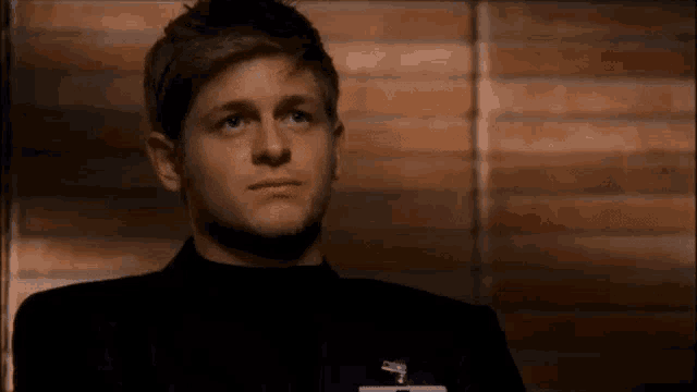 a man in a black suit is sitting in front of a brick wall and looking up .
