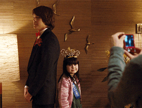 a man in a suit stands next to a little girl wearing a crown and a blue ribbon with the letter f on it