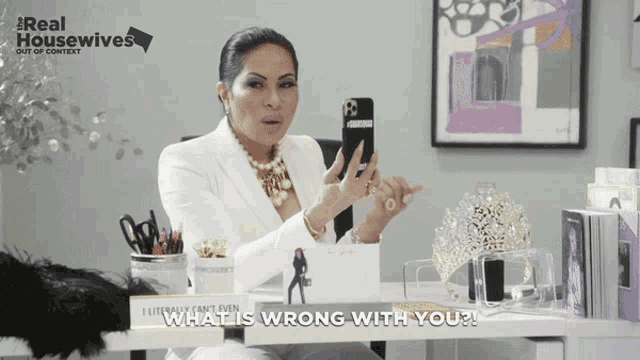 a woman sitting at a desk holding a cell phone with the words " what is wrong with you " written below her