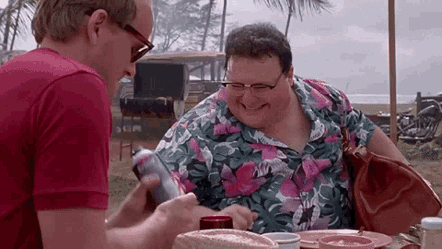 two men are sitting at a table and one is holding a can of soda