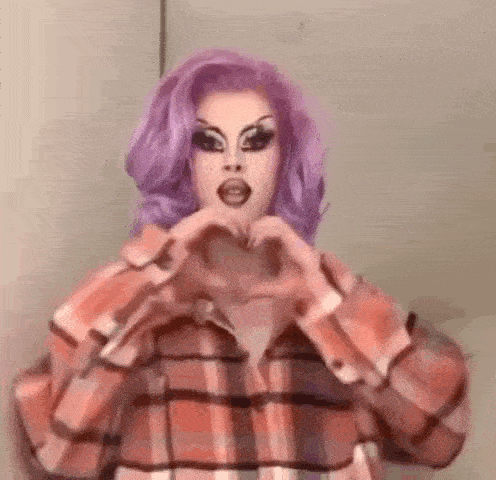 a drag queen with purple hair and a plaid shirt is making a heart with her hands .