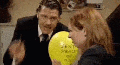 a man in a suit and tie is blowing up a yellow balloon that says `` jen 's peace of mind '' .