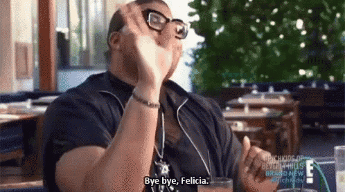 a man wearing glasses is sitting at a table with a drink in his hand and says `` bye bye felicia ''