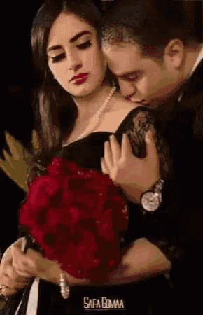a man is kissing a woman on the cheek while holding a bouquet of red roses .